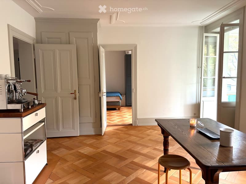 Bright, open-plan living and dining area with wooden herringbone flooring, large windows, a wooden dining table with a stool, a coffee machine on a counter, and a visible bedroom with a bed through an open doorway.