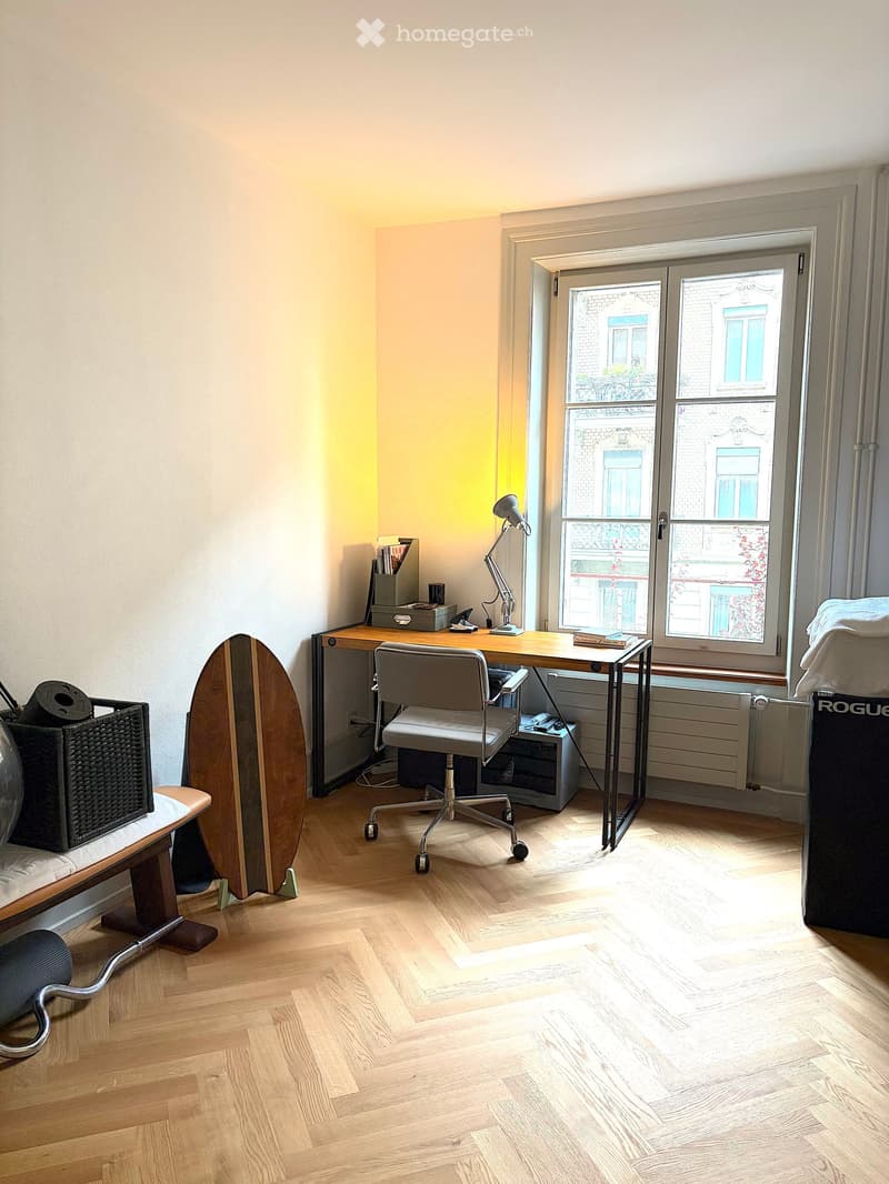 A well-lit room with wooden flooring, a desk with a chair, a lamp, a surfboard leaning against the wall, and a small table with a basket on it.