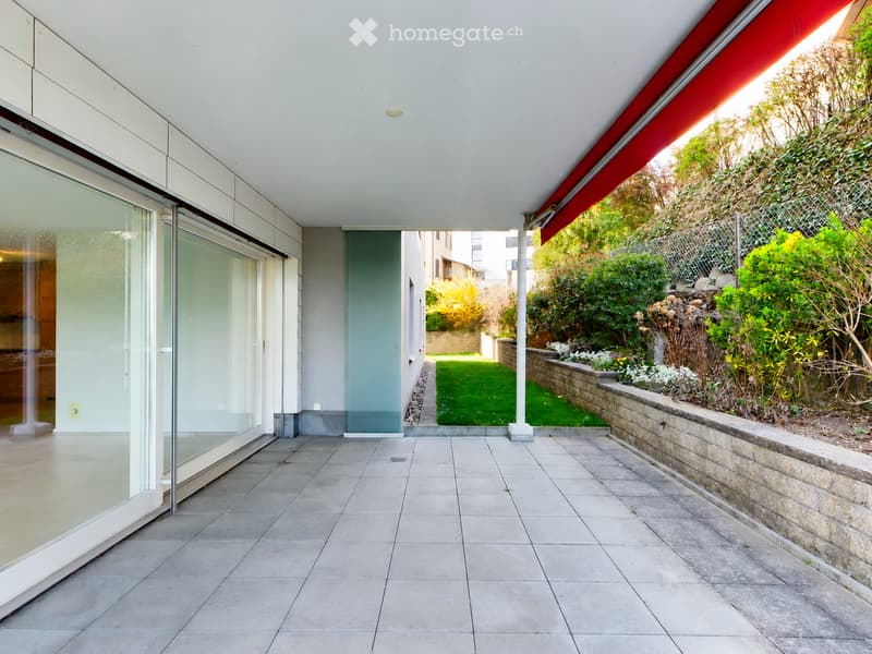 Modern outdoor patio with tiled flooring, glass sliding doors leading to the interior, a raised stone border, and a well-maintained garden with green grass and various plants.