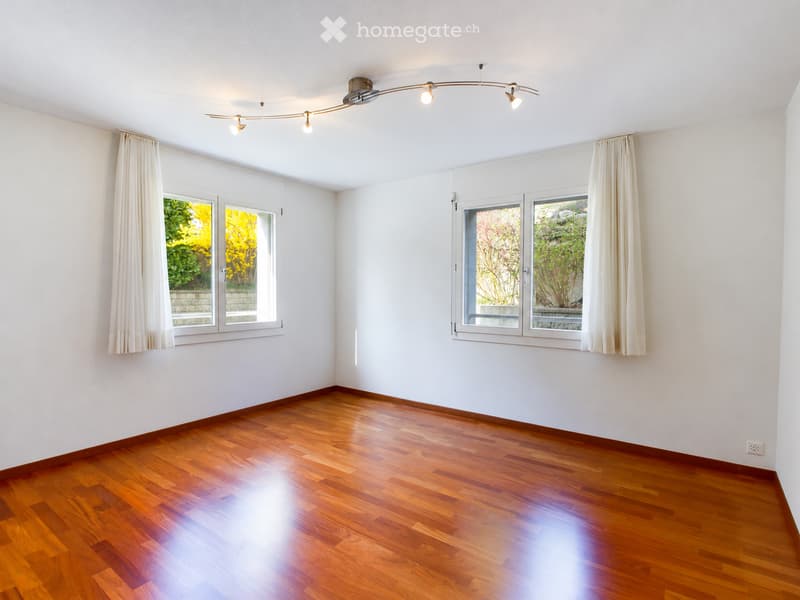 Empty room with wooden flooring, white walls, large windows with white curtains, and a modern track light fixture on the ceiling.