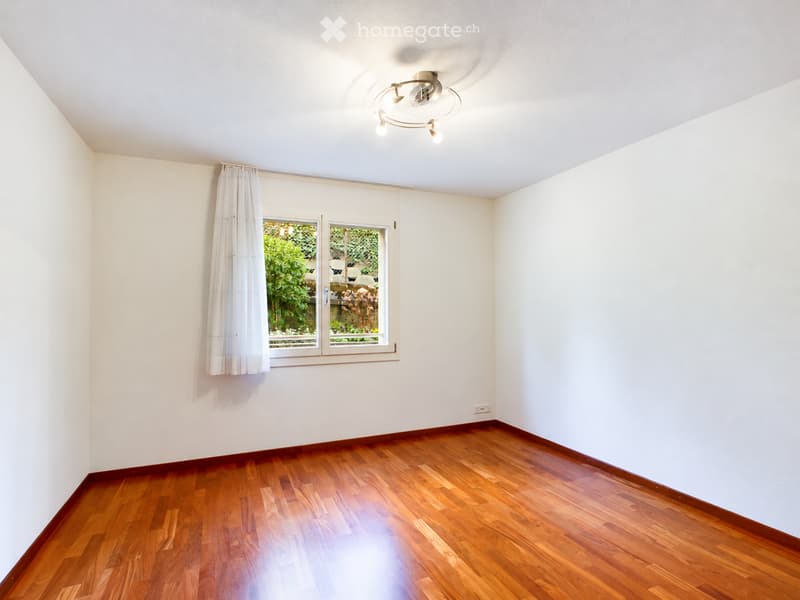 Empty room with wooden flooring, white walls, and a window with white curtains. The ceiling features a modern light fixture.