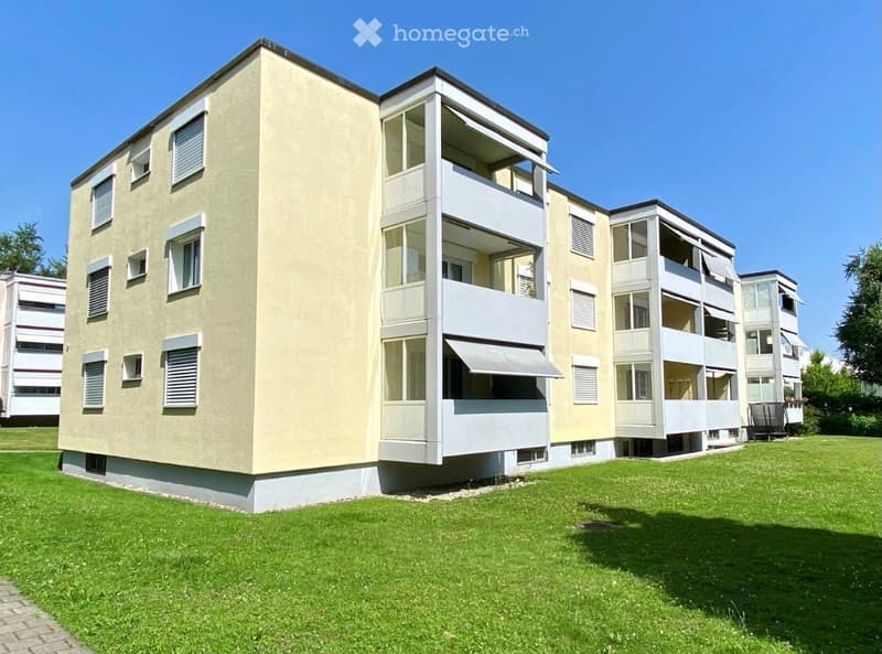 Modern multi-story residential building with balconies, large windows, and a well-maintained lawn in front.