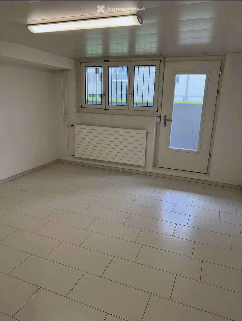 Empty room with tiled floor, white walls, a radiator, and a door with a window. The room has three windows with metal bars and a fluorescent ceiling light.