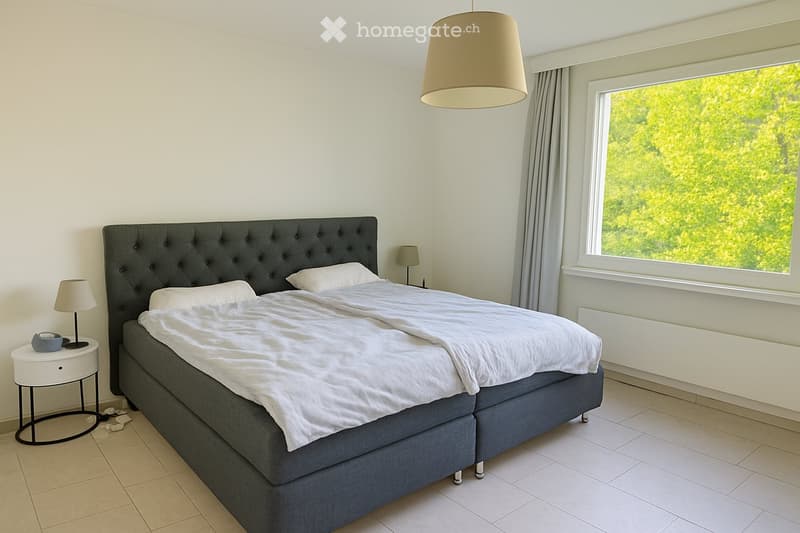 Modern bedroom with a large bed, white bedding, nightstand with lamp, and large window with view of greenery.