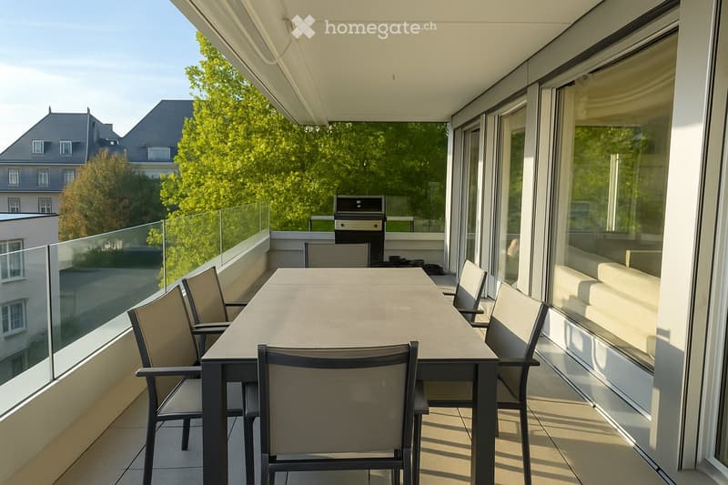 Modern balcony with a glass railing, a dining table with four chairs, and a grill in the background.
