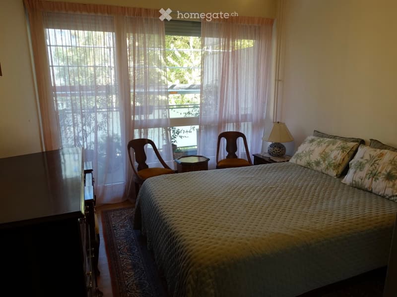 Cozy bedroom with a large bed, patterned pillows, a nightstand with a lamp, two wooden chairs, a small table, and sheer curtains covering the windows.