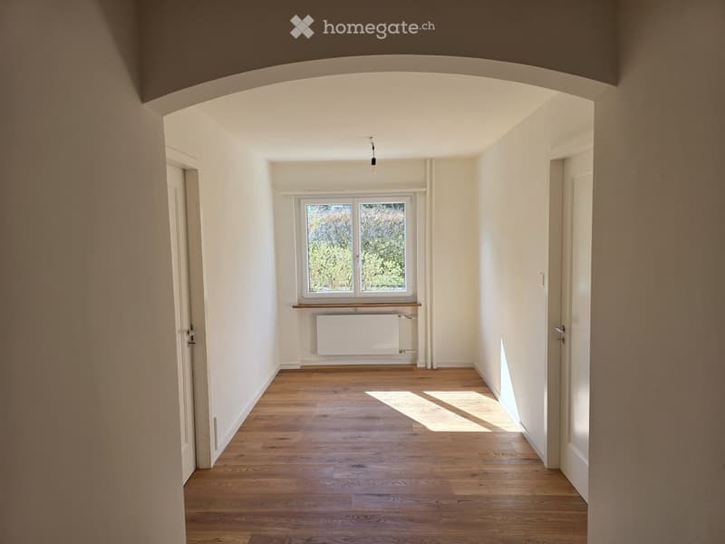 Empty hallway with wooden flooring, white walls, and a window at the end. Natural light streams in, casting shadows on the floor.
