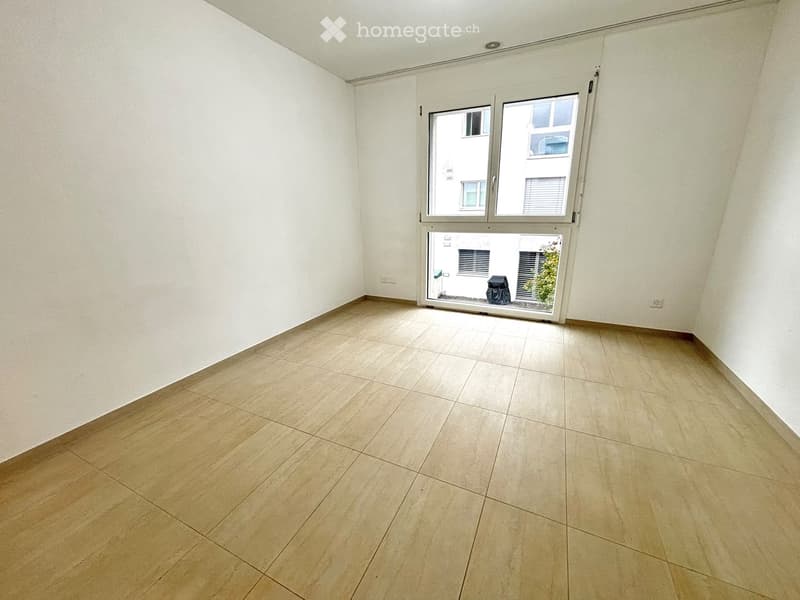 Empty room with light-colored tiled flooring and white walls, featuring a large window with a view of an adjacent building.