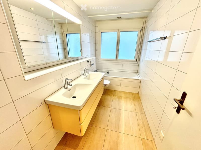 Modern bathroom with white tiles, a double sink, a bathtub, and a large mirror. The floor is tiled in a light wood finish.