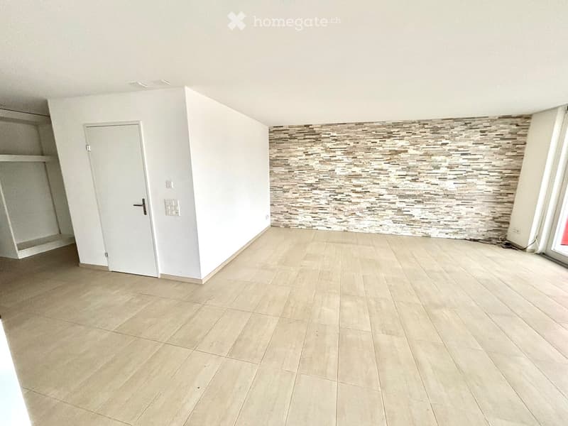 Empty room with light-colored tiled flooring, white walls, and a decorative accent wall with a mix of brown and beige tiles. A white door is visible on the left side.