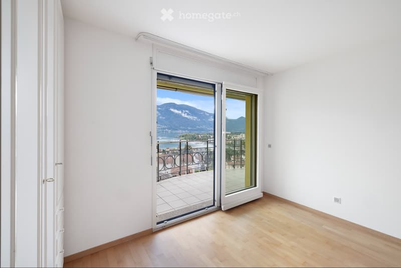 Empty room with white walls, wooden floor, sliding glass doors leading to a balcony with a view of the lake and mountains.