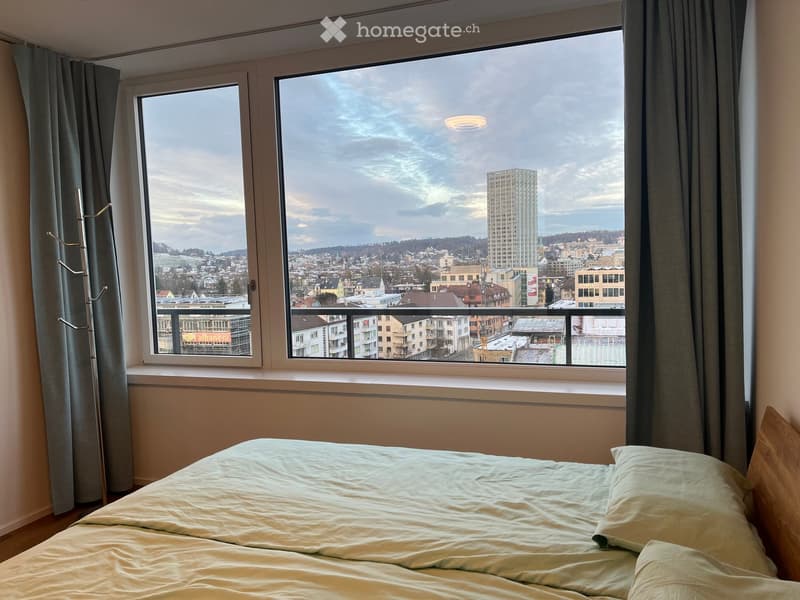 Bedroom with a large window offering a cityscape view, featuring a neatly made bed and curtains.