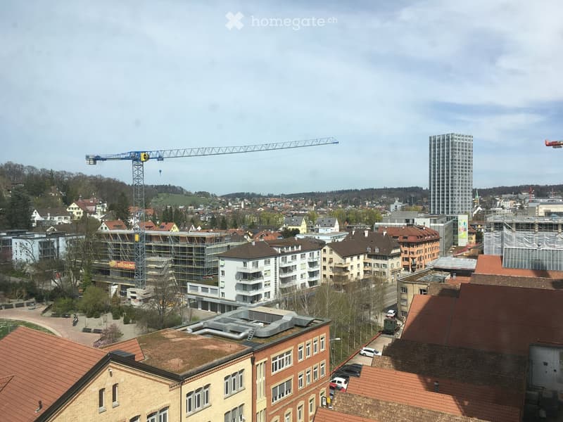 A panoramic view of a cityscape with multiple buildings, including residential and commercial structures, a construction crane, and a tall skyscraper in the background.