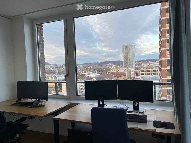 A modern, well-lit office space with large windows offering a cityscape view. The room features two desks with dual monitors, a comfortable office chair, and essential computer peripherals.