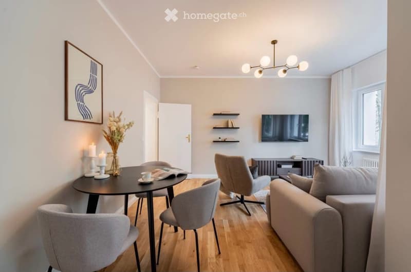 Modern living room with light-colored furniture, a sofa, a dining table with chairs, a television, shelves, and decorative items including a vase with flowers and candles.