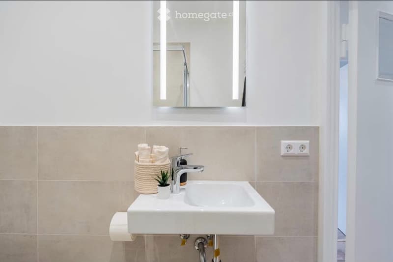 Modern bathroom with a white sink, chrome faucet, large mirror, and a woven basket holding towels and a small plant. Beige tiled walls and a white toilet paper holder are visible.