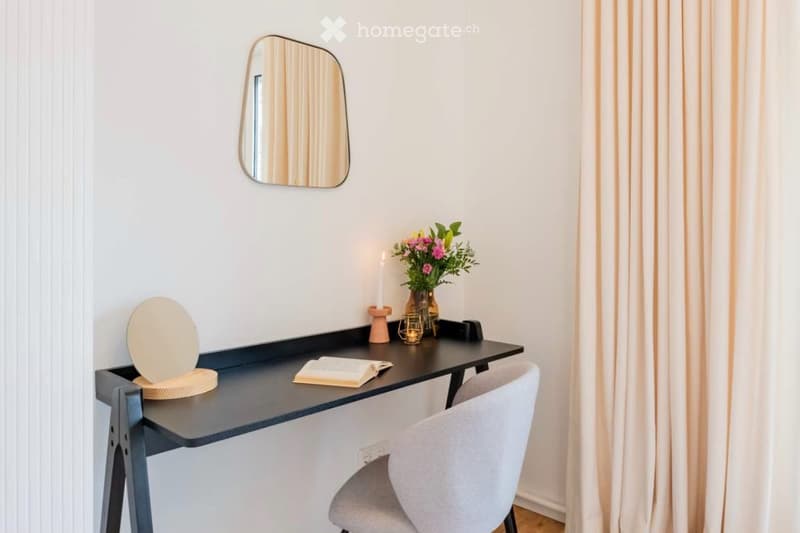 A modern, minimalist desk area with a sleek black desk, grey chair, round mirror, vase with flowers, candle, and a book.