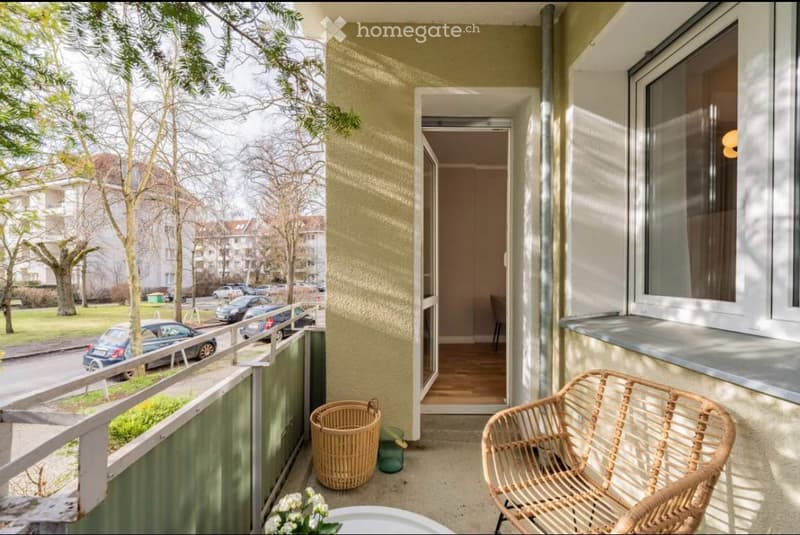 Balcony with a wicker chair, a wicker basket, and a small table with flowers, overlooking a street with parked cars and trees.