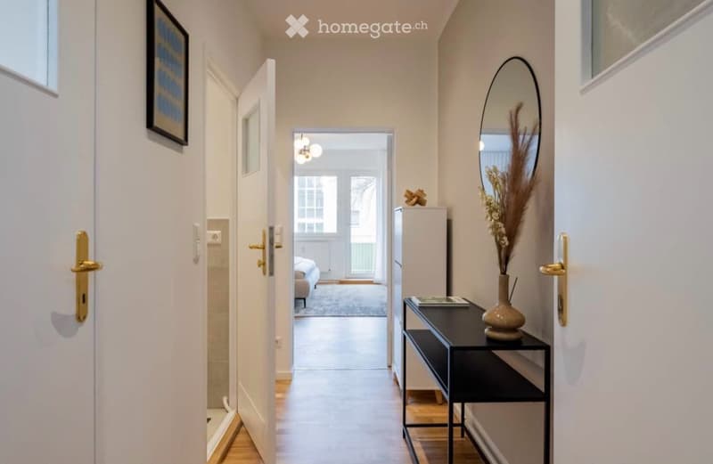 Modern hallway with white walls, wooden flooring, and a small black console table with a vase and mirror. Open doorway leads to another room.