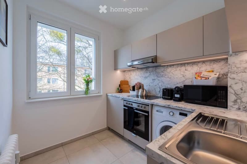 Modern kitchen with white cabinets, marble countertops, stainless steel appliances, and a window view of the street.