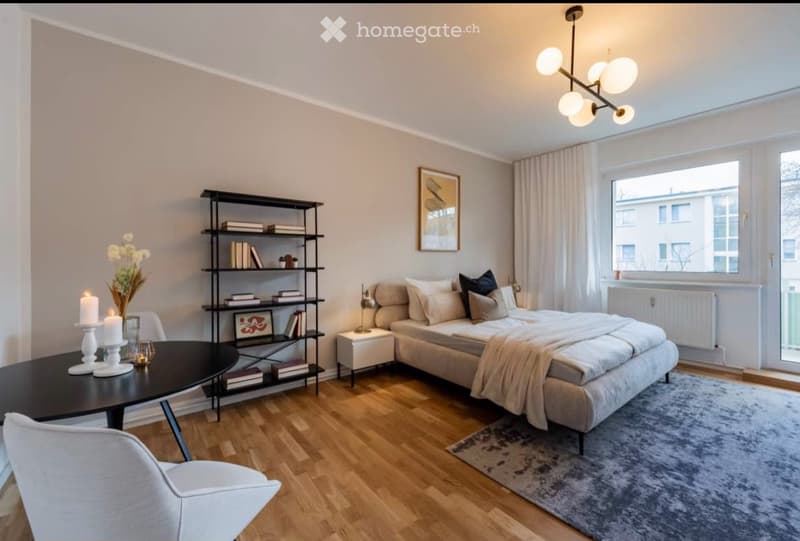 Modern bedroom with a large bed, nightstand, round table, chair, bookshelf, and decorative items including candles and a vase.