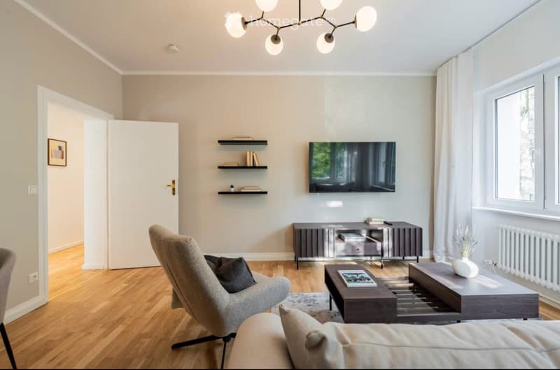 Modern living room with light-colored walls, wooden flooring, a grey sofa, a coffee table, a TV mounted on the wall, and a sleek entertainment unit.