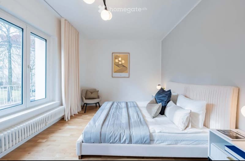 Modern bedroom with a large bed, nightstand, armchair, and artwork on the wall. Large windows with curtains provide natural light.