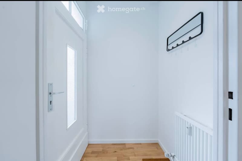A narrow, white-walled hallway with wooden flooring, a white door on the left, and a small mirror with hooks on the right wall.