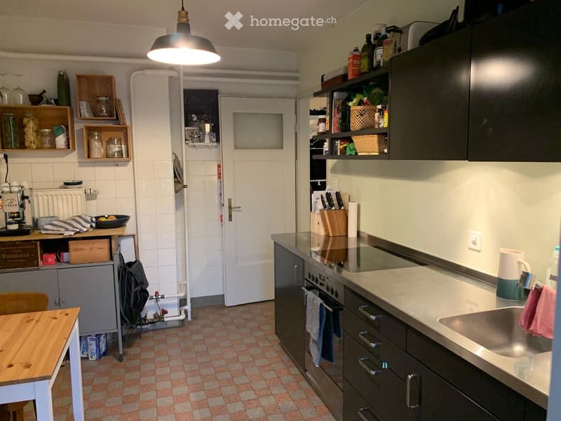 The kitchen features a wooden table, stainless steel oven, sink, and various cabinets. Shelves hold jars and kitchen items, and a coffee machine is on the counter.