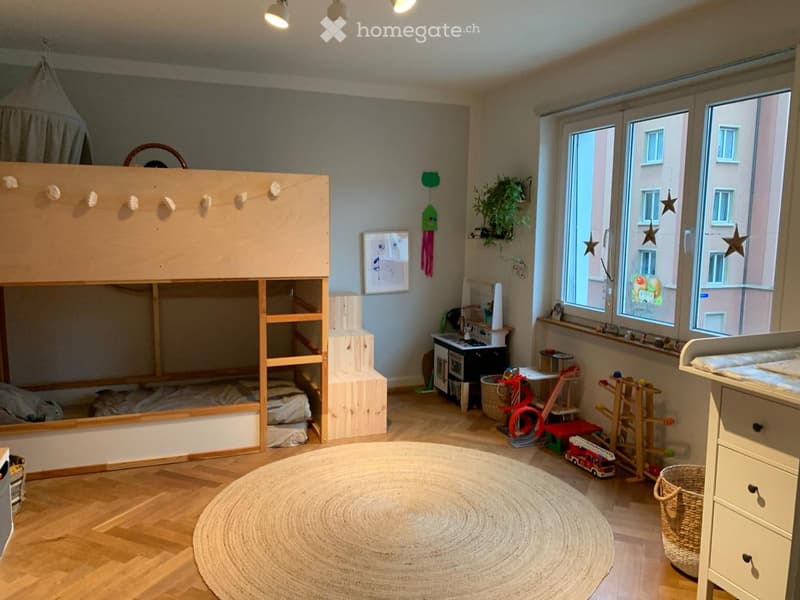A well-lit children's bedroom with a lofted wooden bed, various toys, a round rug, and a view of the city through large windows.