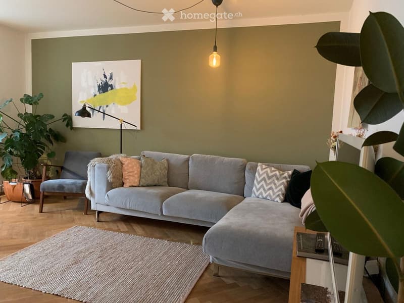 Modern living room with a grey sectional sofa, throw pillows, a wooden side table, a pendant light, a large painting, and a potted plant.