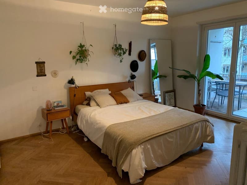 Cozy bedroom with a large bed, wooden nightstand, table lamp, mirror, and potted plants. A wicker hanging lamp adds warm lighting, and a sliding glass door leads to a balcony area.