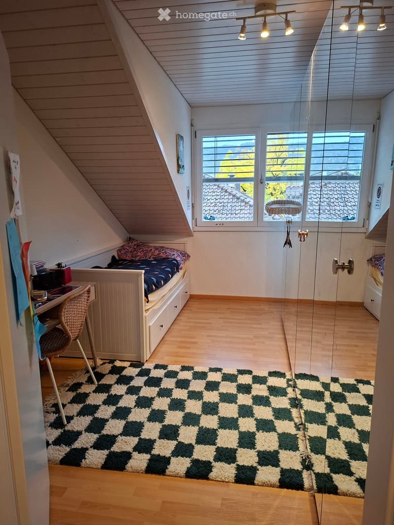 Cozy attic bedroom with wooden floors, slanted ceilings, and large windows. Features a built-in bed with drawers, a desk with a chair, and a checkered rug.