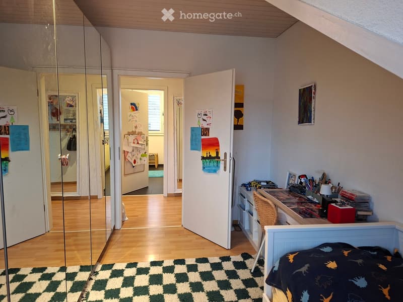 A furnished bedroom with a bed, desk, chair, mirror, and storage unit. The room features wooden flooring, a checkered rug, and several doors leading to other areas.
