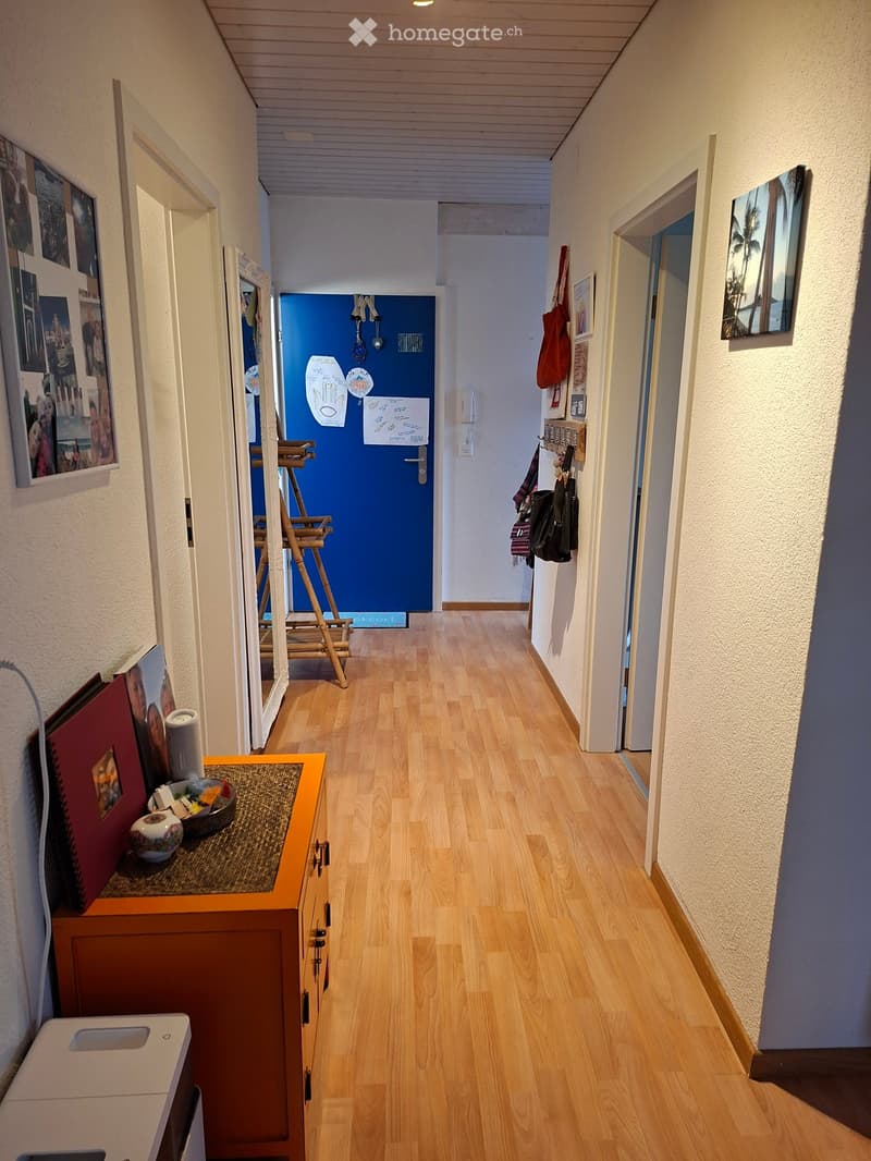 A well-lit hallway with wooden flooring, white walls, and several doors. There are framed pictures on the walls, a small table with decorative items, and a coat rack with bags and coats.