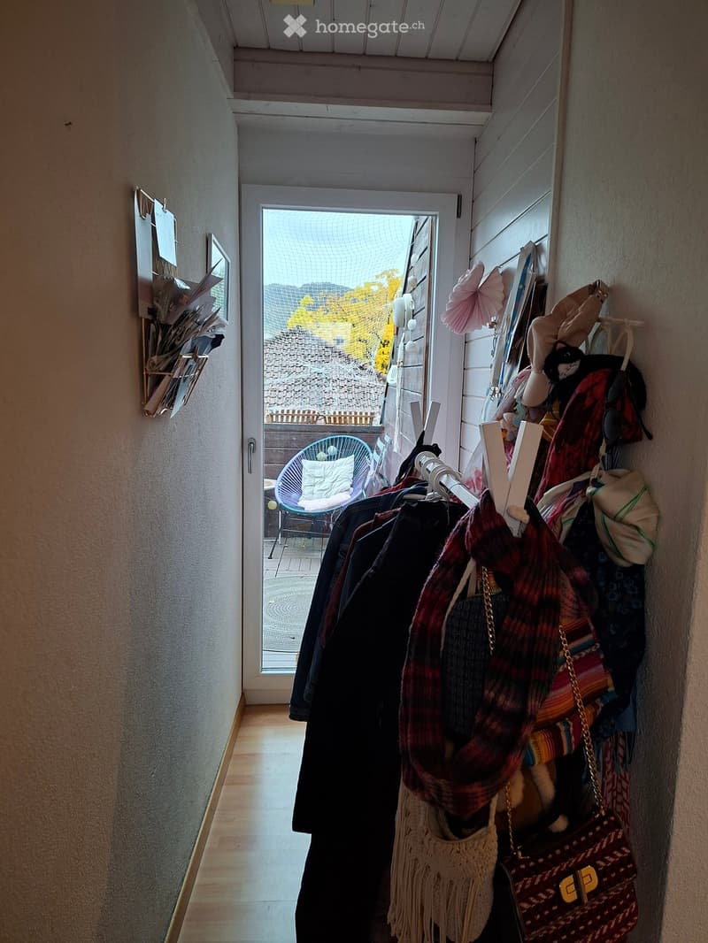 A narrow hallway with wooden flooring, light-colored walls, and a coat rack holding various clothing items, bags, and a cat. An open door leads to a balcony area with a chair and a view of the outside.