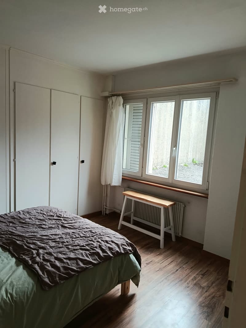 Bedroom with wooden flooring, a bed with green and brown bedding, a small white stool, a radiator, and a white wardrobe.