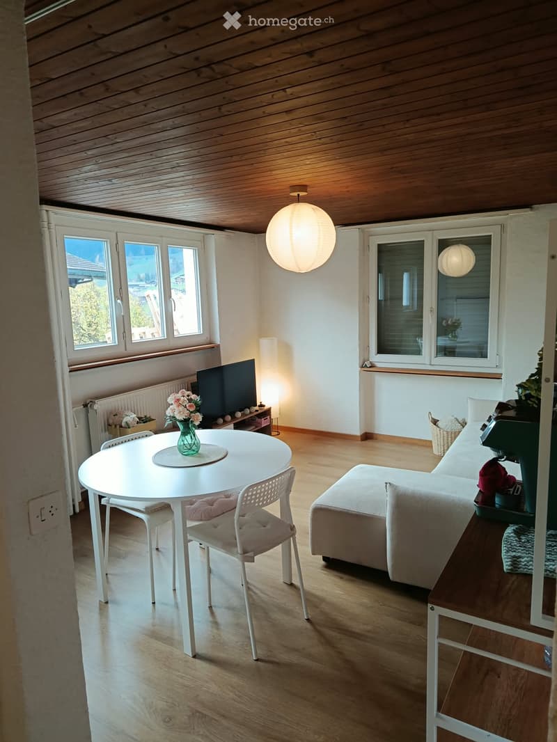 A modern, well-lit living room with wooden flooring, white walls, and large windows. Features include a white sofa, a small round table with chairs, a television, and decorative vases.