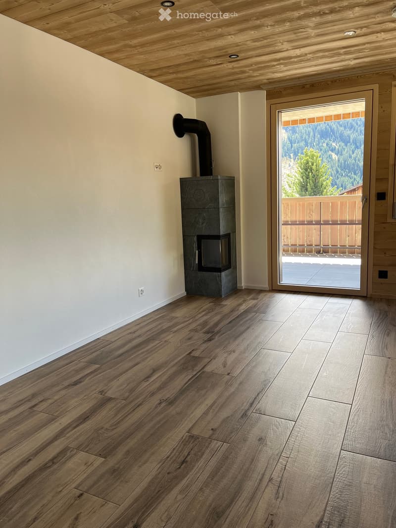 Empty room with wooden flooring, white walls, and a modern fireplace. A large sliding glass door opens to a balcony with a scenic mountain view.
