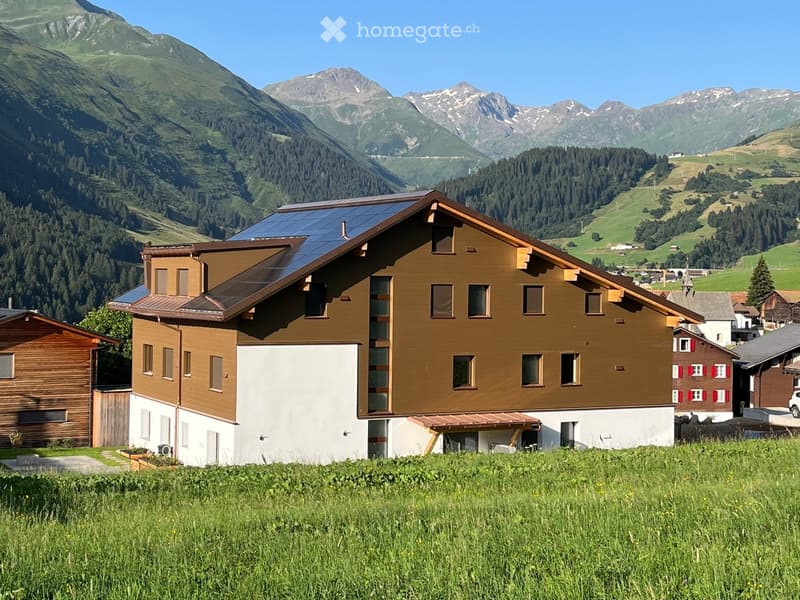 A two-story wooden house with solar panels on the roof, surrounded by green fields and mountains.