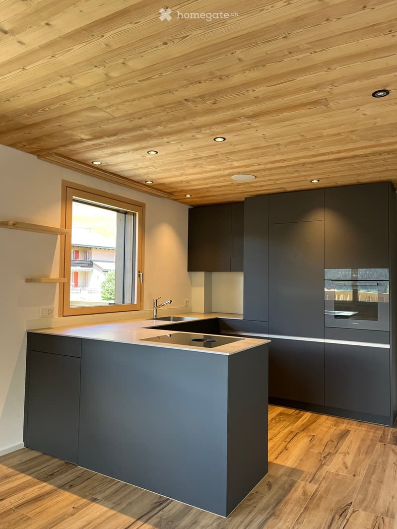 Modern kitchen with dark grey cabinets, a central island, built-in oven, and a large window offering a view of the outside.