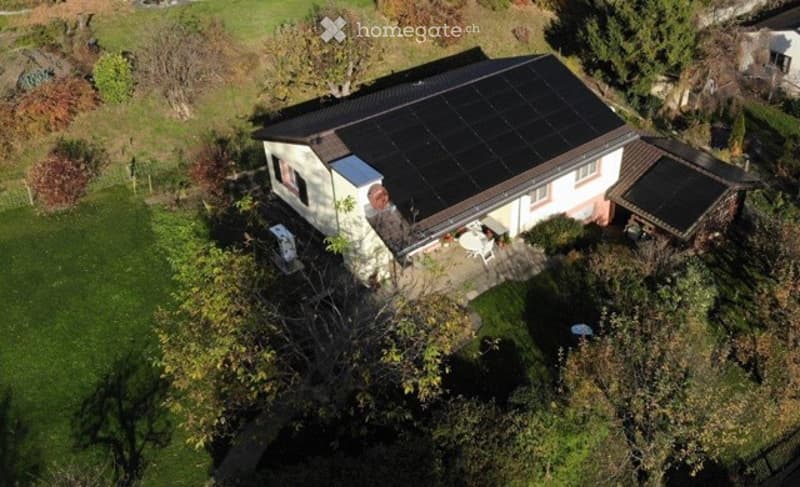 Aerial view of a two-story house with solar panels on the roof, surrounded by a well-maintained lawn and trees. A small patio area with outdoor furniture is visible near the house.