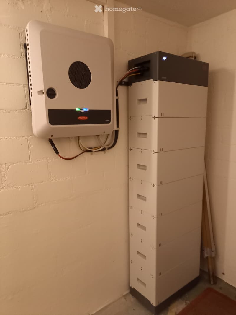 A room with a white brick wall and two energy storage units: a wall-mounted inverter and a stacked battery unit.
