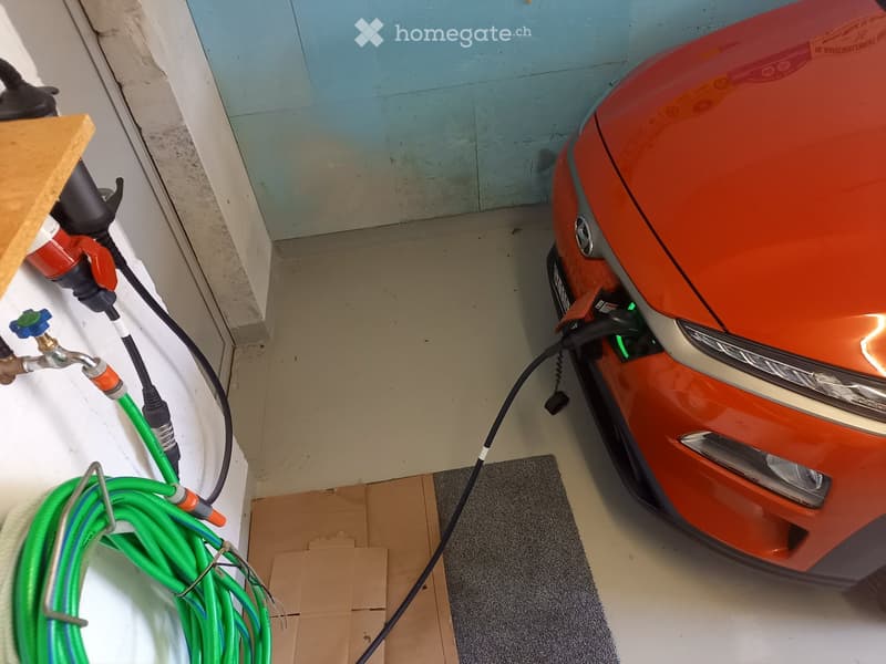 A red car is plugged into a charging station inside a garage. Green hoses and cables are visible on the floor.