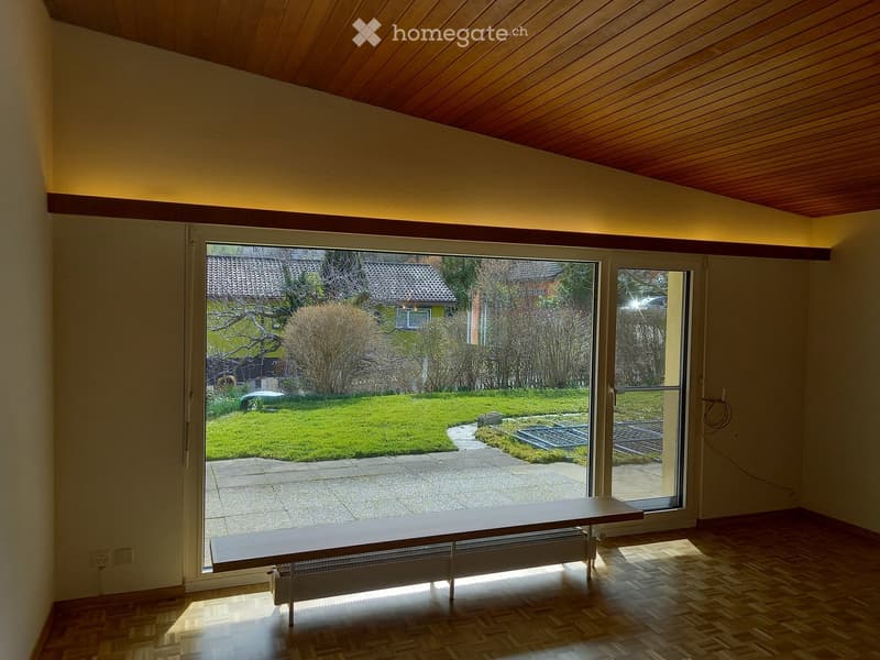 Bright, empty room with wooden flooring and a slanted wooden ceiling. Large glass doors open to a garden view, with a bench placed just inside the doorway.