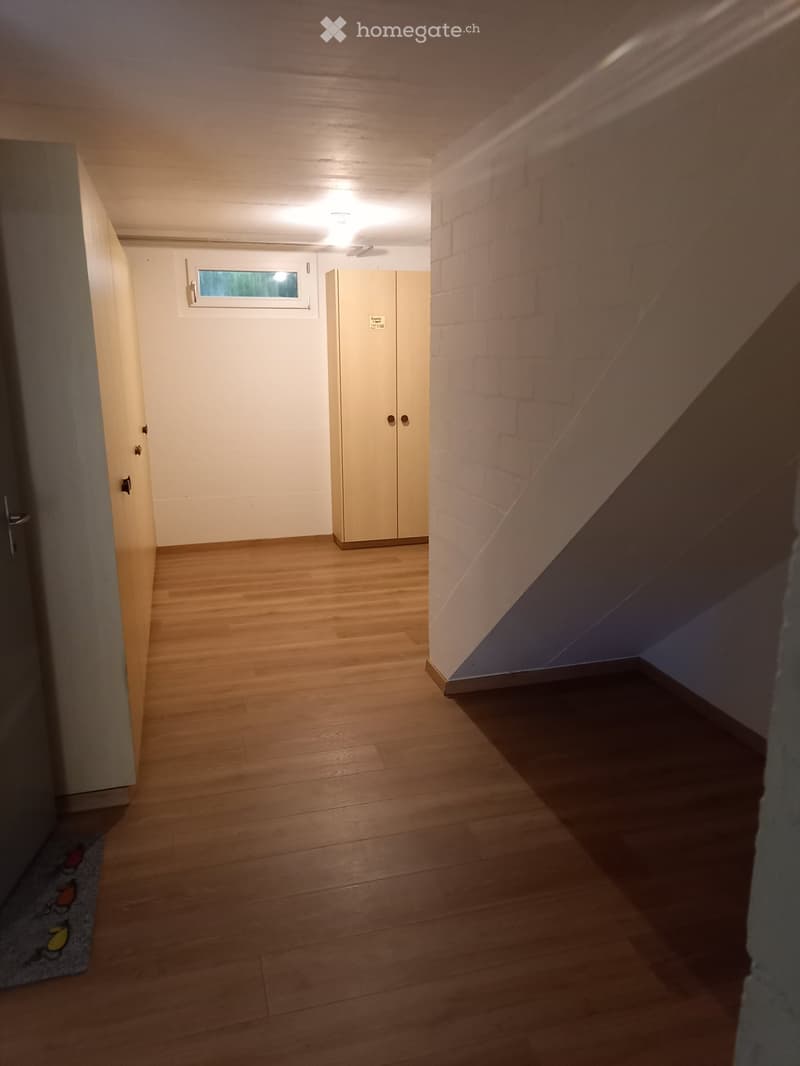 A narrow hallway with wooden flooring, white walls, and a small window. A staircase leads upwards, and a closet is visible on the right side.