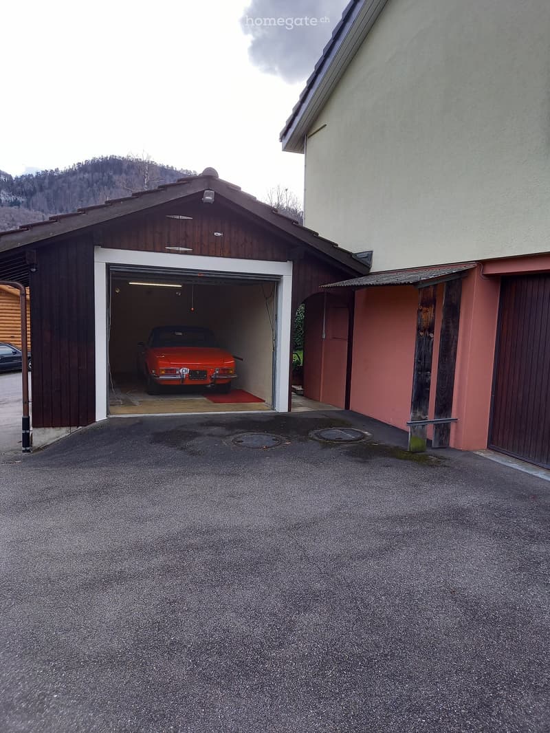 A single-car garage with a red vintage car inside, attached to a larger building with a sloped roof and wooden siding.