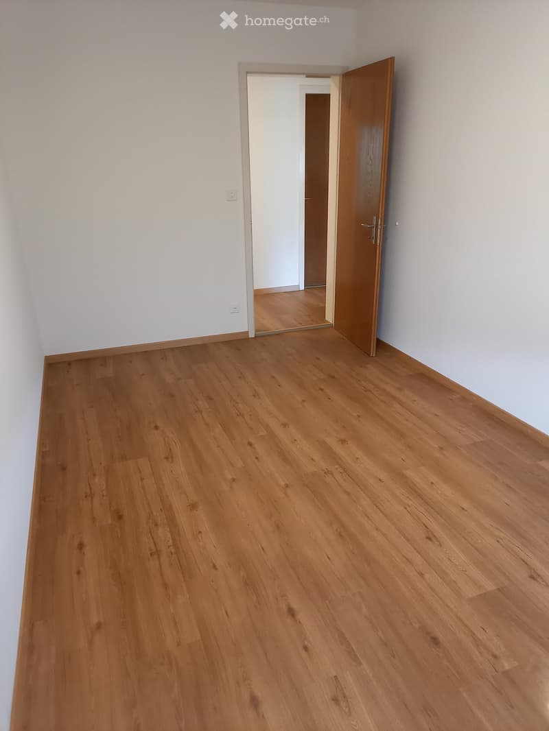 Empty room with light wooden flooring and white walls, featuring an open doorway leading to another room.