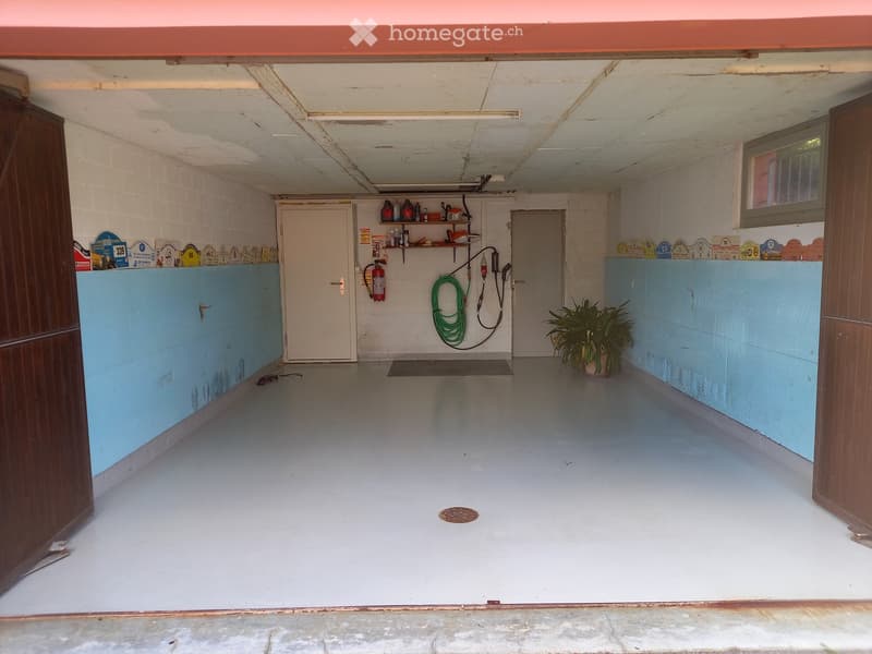 Empty garage with white brick walls, blue lower panels, and a concrete floor. Features include a fire extinguisher, shelves with various items, a green hose, and a potted plant.