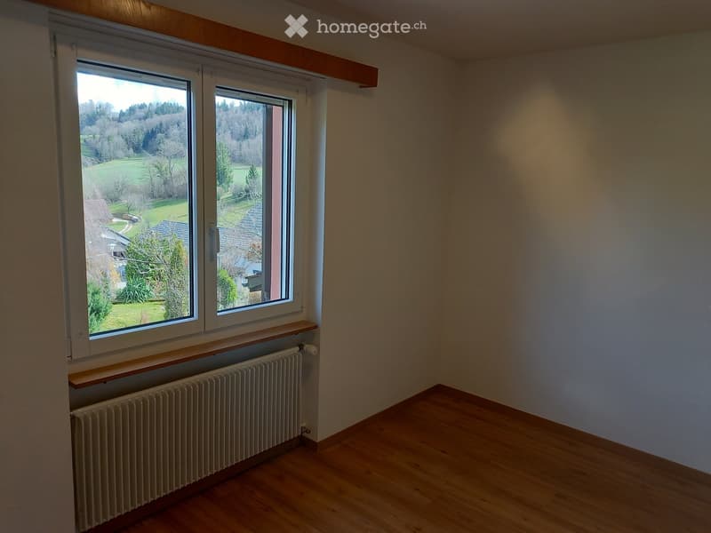 Empty room with wooden flooring, large windows offering a view of a green landscape, radiator, and wooden shelf under the windows.